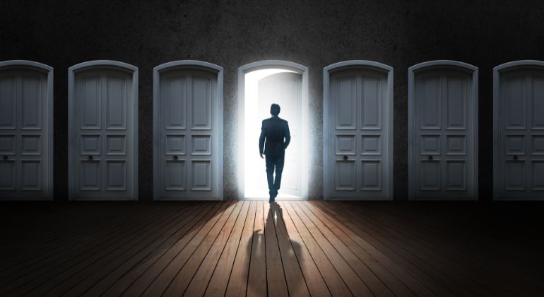 Silhouette of a man walking through an open, glowing door among several closed white doors, symbolizing career choices and future opportunities, representing career astrology predictions in India.
